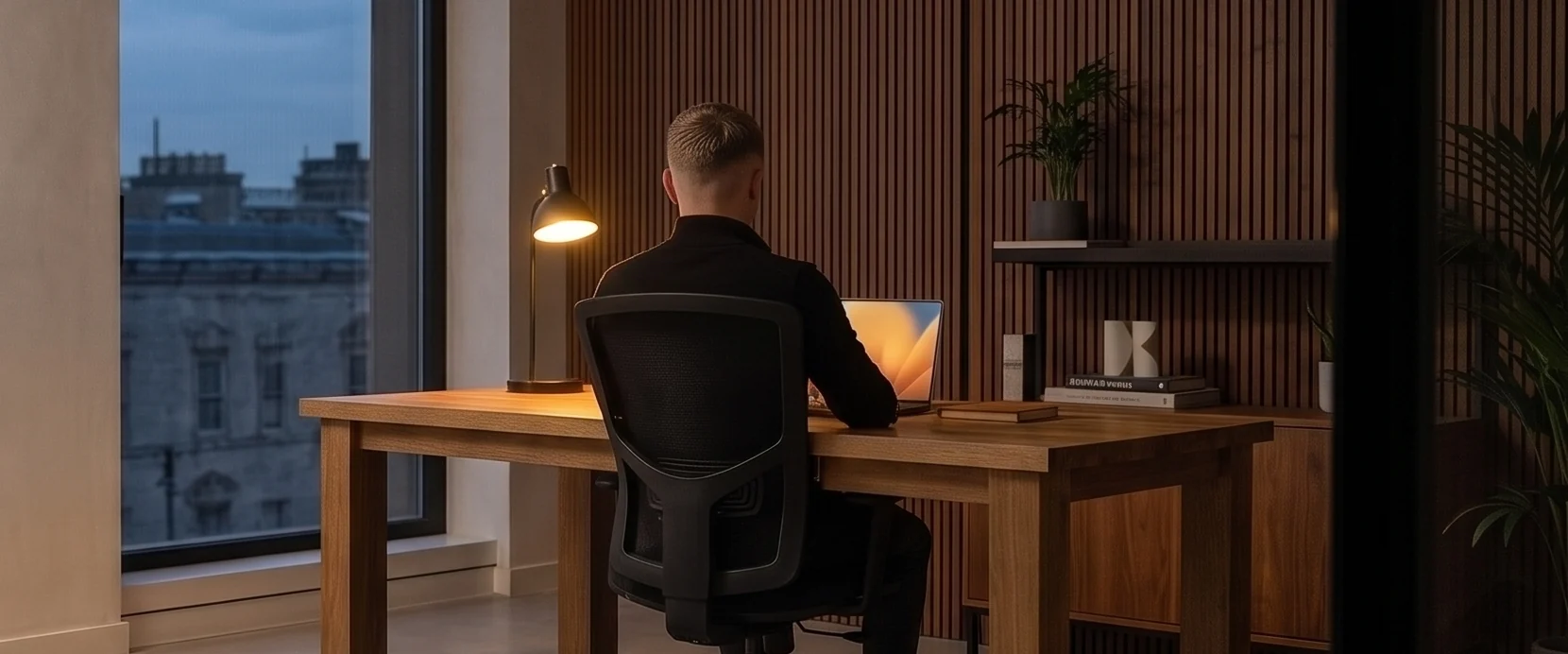 Dominic Wolter arbeitet abends allein an seinem Schreibtisch, die Schreibtischlampe ist die einzige Lichtquelle im Raum, draußen blaue Dämmerung über den Dächern